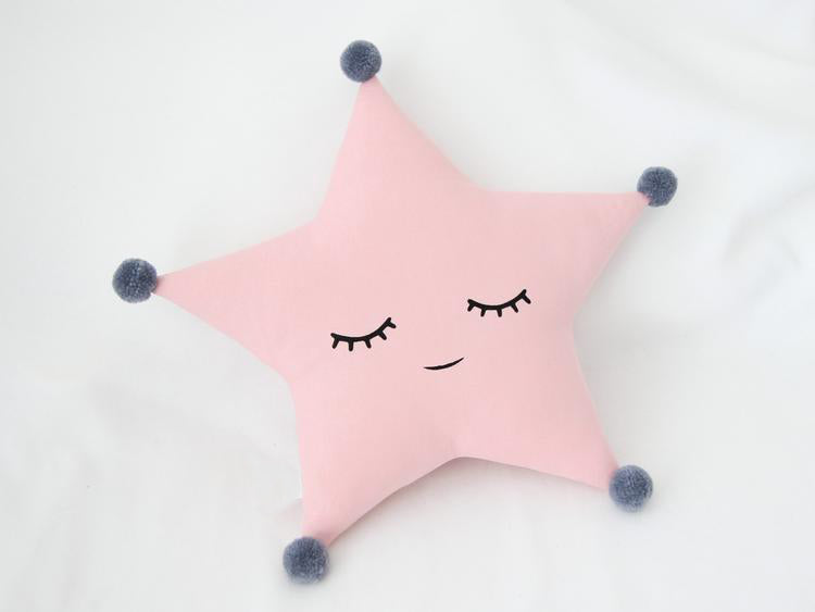 Cute grey star pillow with eyes and pom poms, perfect for adding a touch of whimsy to your home decor.