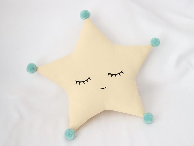 Cute grey star pillow with eyes and pom poms, perfect for adding a touch of whimsy to your home decor.
