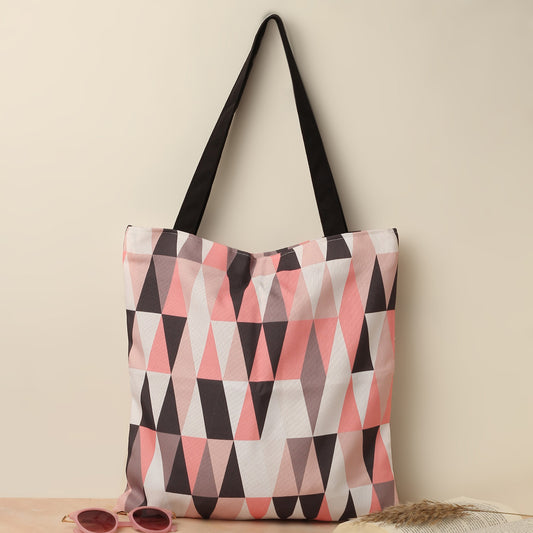 A tote bag with a geometric pattern in shades of pink, black, and white, with black handles placed on a beige surface next to a pair of pink sunglasses and dried grass.