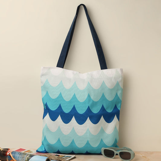 A tote bag with a blue and white wave pattern, navy blue handles, placed on a beige surface next to a pair of teal sunglasses and travel brochures.