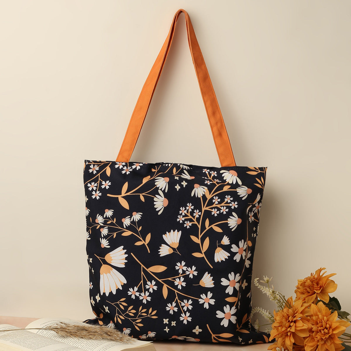 A black floral tote bag with orange straps leaning against a light beige background next to a bunch of orange flowers and an open book.