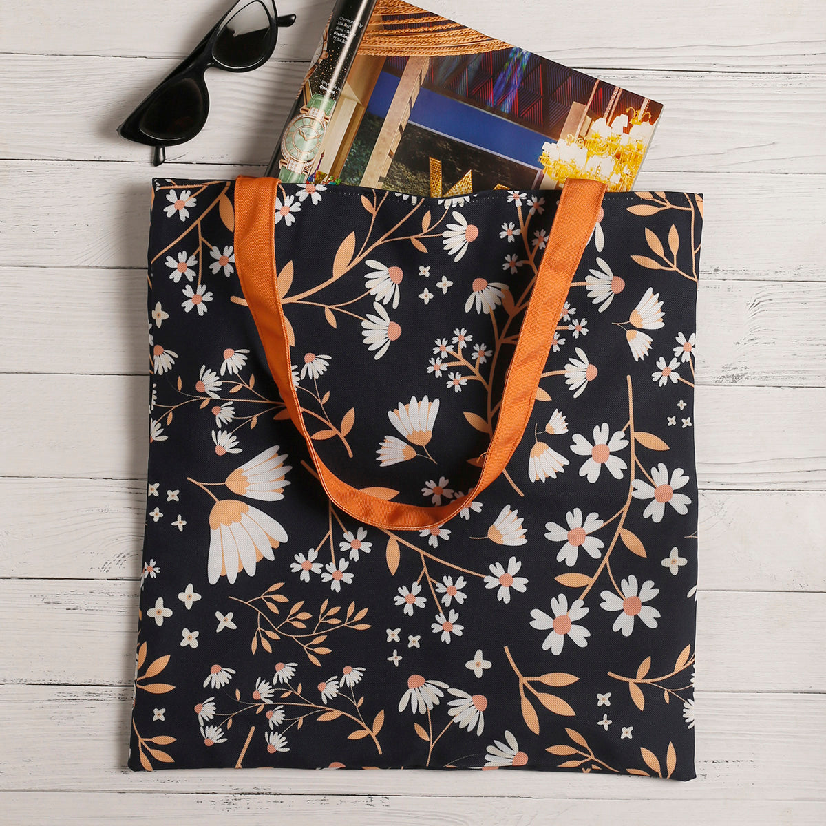 A black tote bag with white and orange floral pattern, orange handles, filled with items such as sunglasses, magazine, and a golden crown, lying on a white wooden surface.