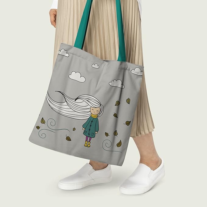 women holding a grey tote bag with a printed design of a girl with long hair, colorful autumn leaves, and clouds. The bag has turquoise handles and the person is wearing white sneakers and beige pleated pants.