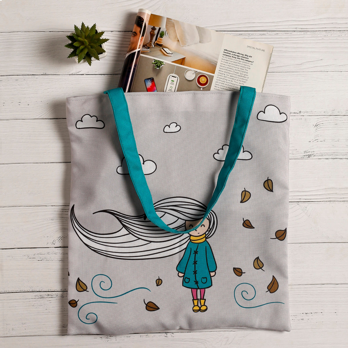 A gray tote bag with a whimsical design of a girl with long flowing hair, clouds, and falling leaves, teal handles, placed on a white wooden surface with a magazine and a succulent plant peeking out from inside.