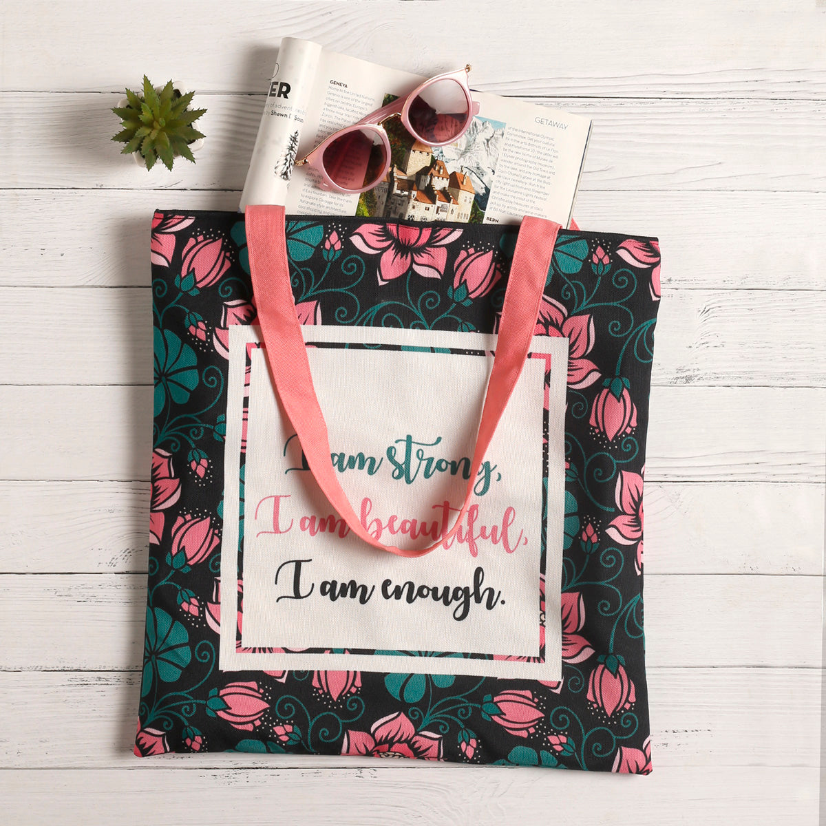 Black tote bag with a quote: ""I am strong, I am beautiful, I am strong"" surrounded by green and pick florals with pink color matching handle with magzine inside & a sunglass nearby put on wooden planks table