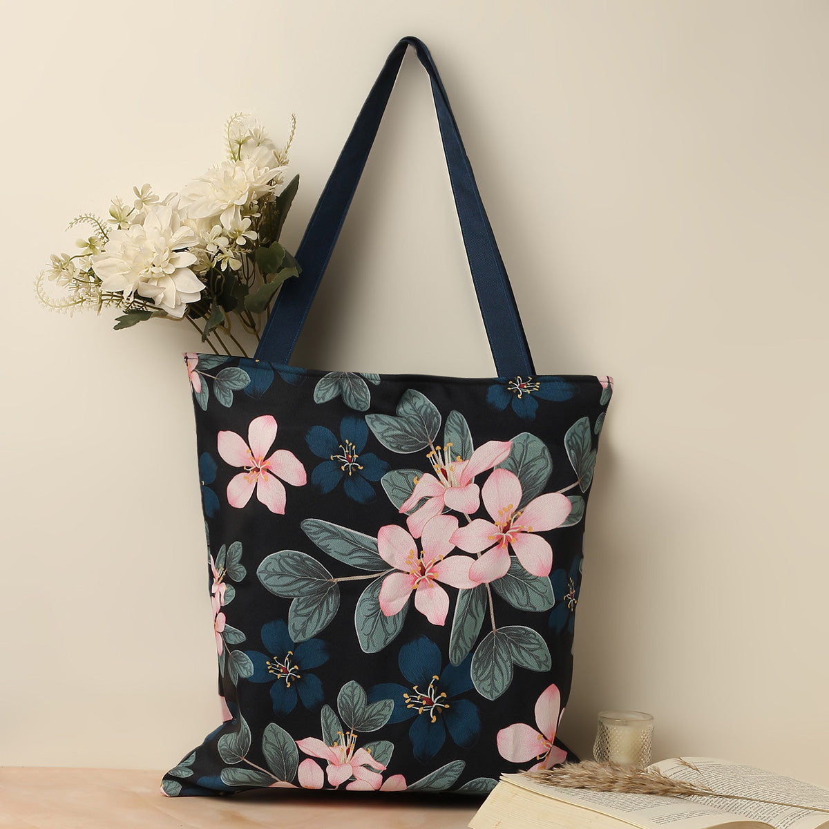 A navy blue tote bag with pink floral print, blue handles, placed next to a bouquet of white flowers and an open book.