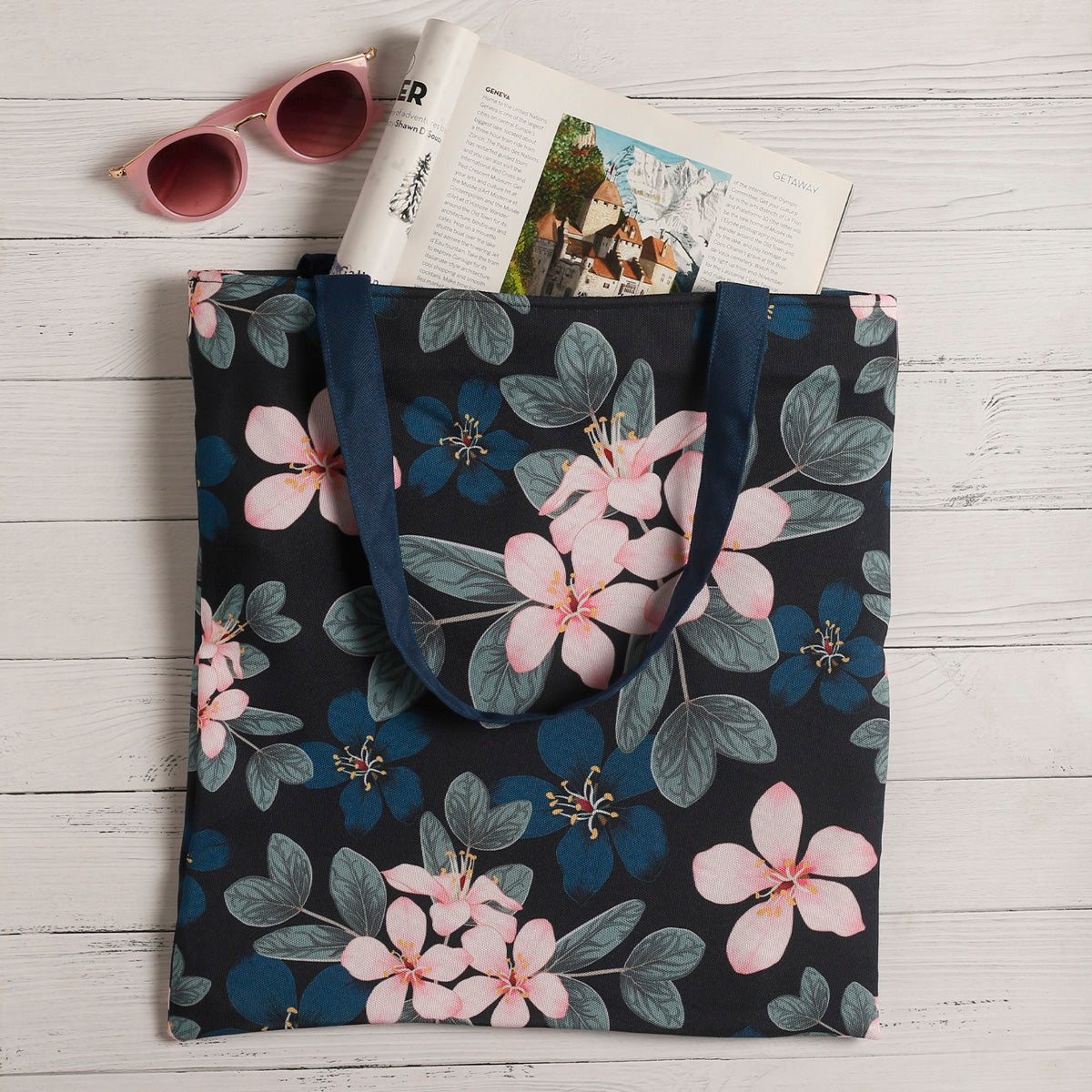 A navy blue tote bag with a pink floral print next to a pair of pink sunglasses and an open magazine on a white wooden surface.