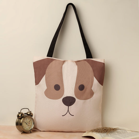 A beige canvas tote bag with a graphic design of a brown and white dog's face, black straps, placed next to an antique alarm clock and an open book with wheat stalks on a cream-colored background.