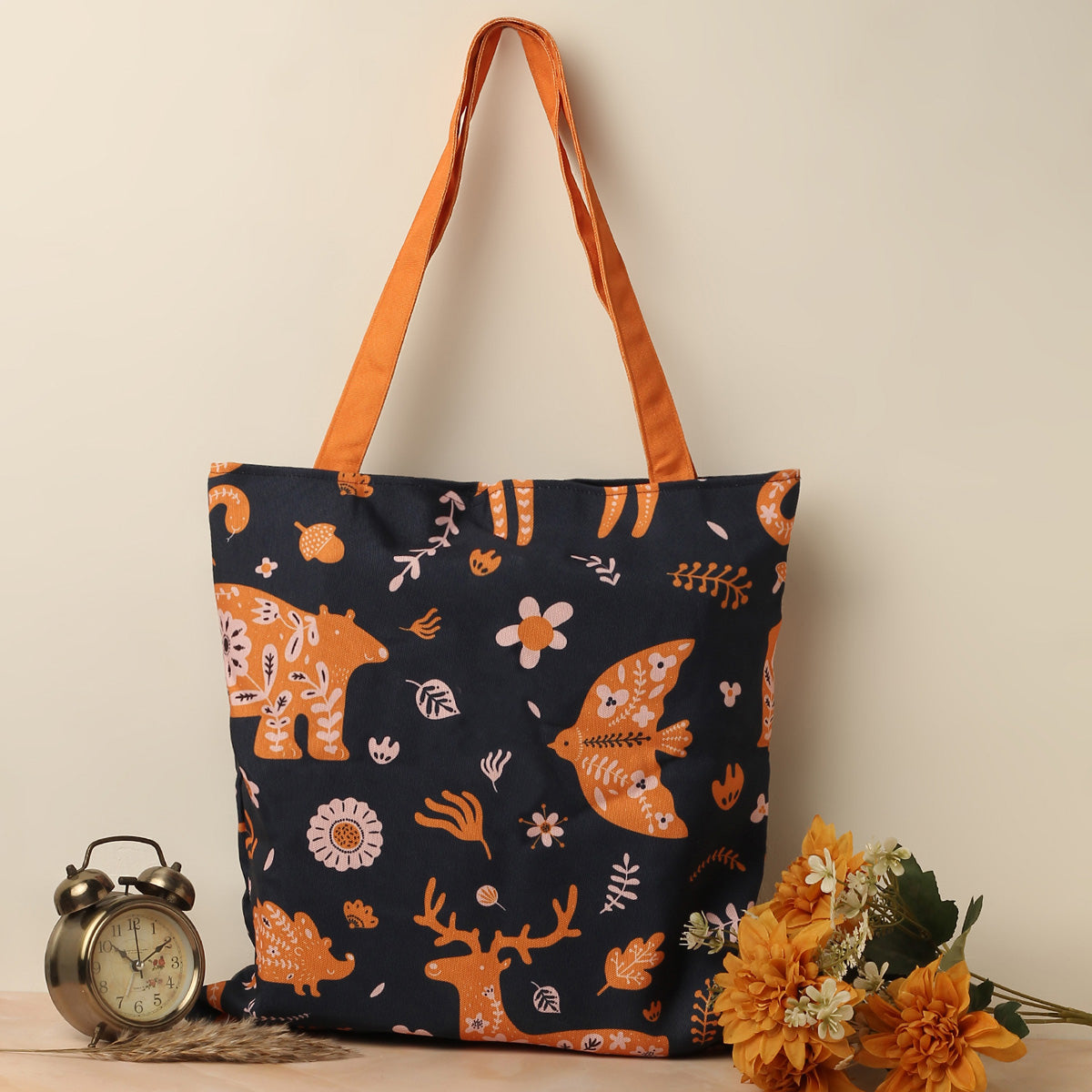 A navy blue tote bag with an orange strap and various orange and white animal and floral designs, accompanied by an alarm clock and a bunch of orange flowers on the side.
