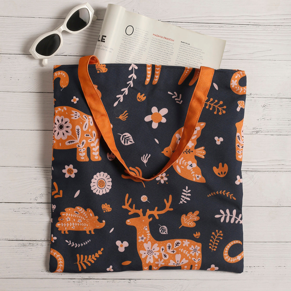 A navy blue tote bag with orange deer and floral patterns, placed on a white wooden surface, accompanied by a pair of white sunglasses and an open magazine.