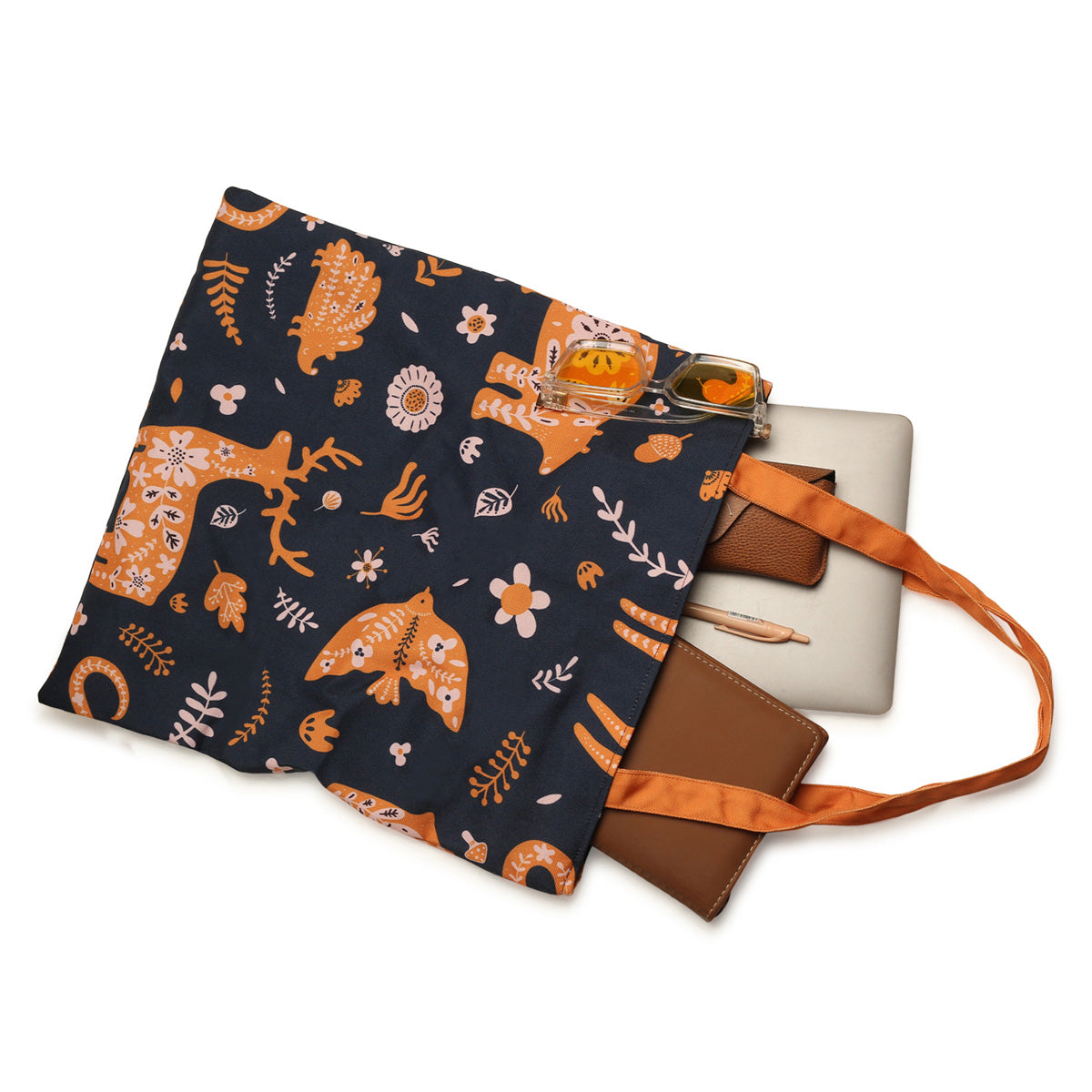 A dark blue tote bag with orange animal and plant prints, an orange strap, and brown leather accents, containing a laptop, a notebook, a pen, and a pair of sunglasses.