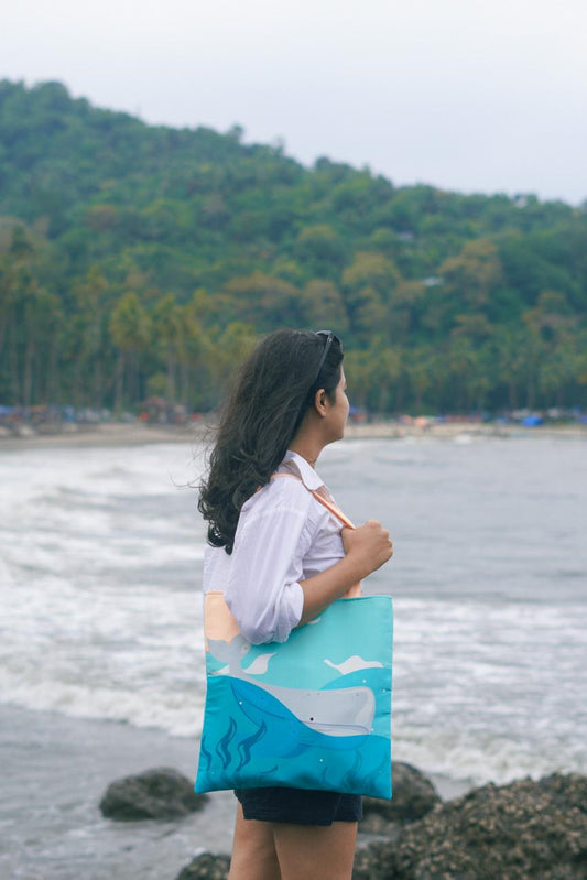 A woman standing on a beach with her back to the camera, holding a tote bag with a whale design, looking out at the ocean and forested hills in the background.