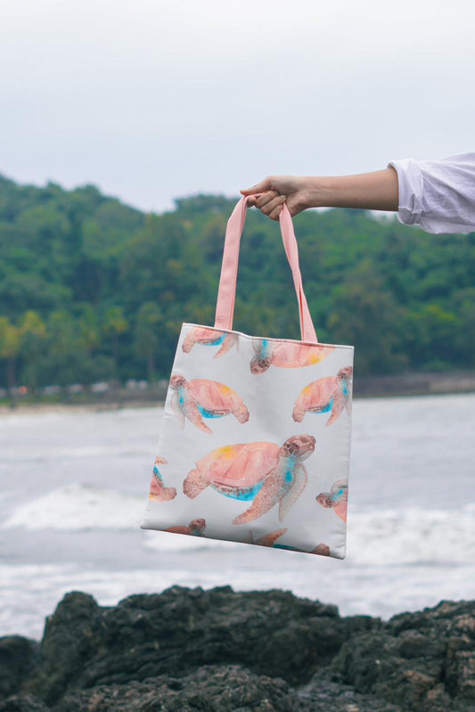 Women holding a tote bag featuring a beautiful watercolor turtle pattern in pink and teal, behind  it these is a river with forest and blue sky above and stones path below.
