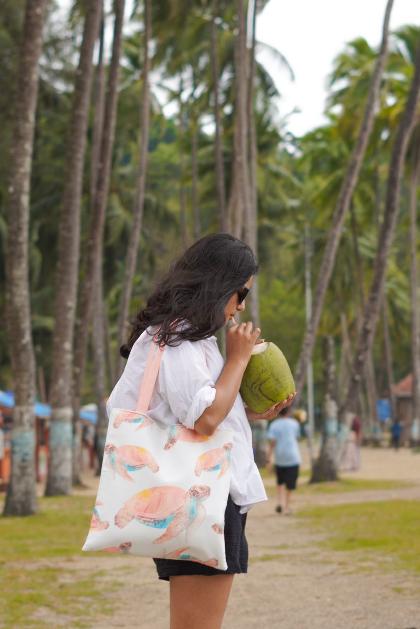 A woman standing on a beach drinking coconut water with coconut trees behind holding a tote bag with cute pastel color turtle prints with color maching handles, enjoying the sunny day by the ocean.