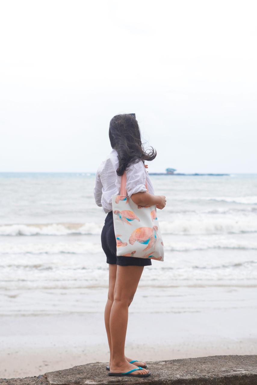 A woman standing on a beach holding a tote bag, enjoying the ocean view