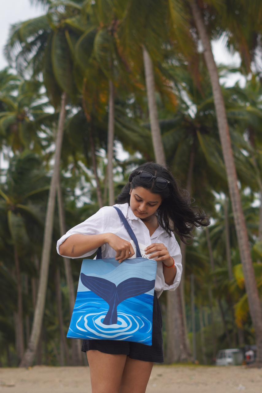 A person standing on a sandy area with palm trees in the background, wearing a white shirt and black shorts, holding a tote bag with a whale tail design against a blue water background. The person's hair is blowing in the wind, and they are wearing sunglasses on top of their head.