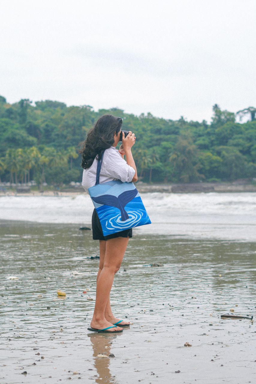 A person standing on a sandy beach holding binoculars to their eyes, wearing a white shirt, black shorts, blue flip-flops, with a shoulder bag featuring a whale tail design. There are palm trees and a hazy sky in the background.