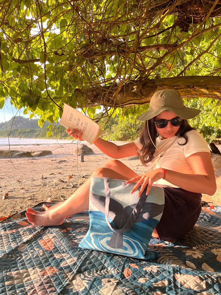 A person sitting under a tree on a beach, reading a book titled "This Is Your Mind on Plants" by Michael Pollan. They are wearing a green cap, white top, and maroon pants, with a blue patterned towel under them. There's a sight of sand and forested hills in the distance, with sunlight filtering through the foliage above.