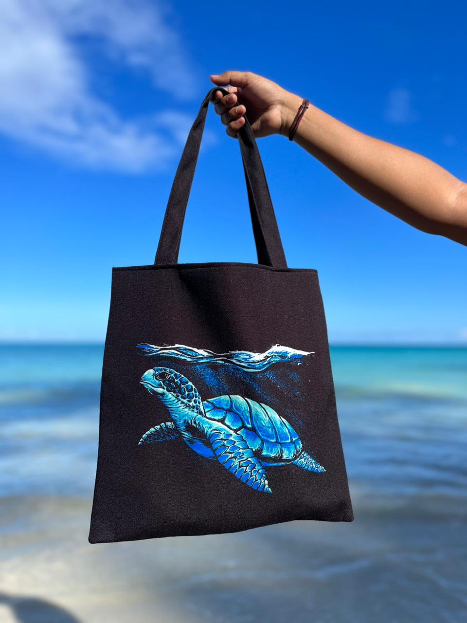 A person is holding a black tote bag with a vibrant blue turtle design on it, with a beach and clear blue sky in the background.