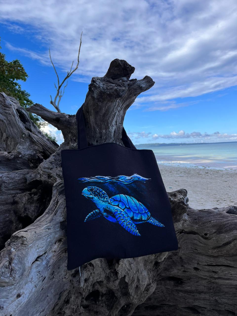  tote bag with a blue turtle design hanging on a driftwood log on a sandy beach, with the ocean and a cloudy sky in the background.
